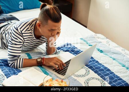 Jungen Mann, der im Schlafsaal auf dem Bett liegt, Kartoffelchips isst, Buch anschaut und sich ein Lernvideo auf dem Laptop ansieht Stockfoto