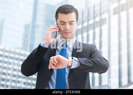 Geschäftsmann, der seine Uhr ansieht, während er anruft, um die Zeit zu überprüfen Handy mit ernsthafteren Gesicht Stockfoto