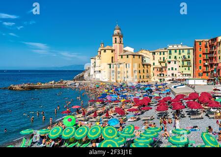 Menschen unter Sonnenschirmen am öffentlichen Strand an sonnigen Tagen als alte bunte Kirche im Hintergrund in Camogli, Italien. Stockfoto