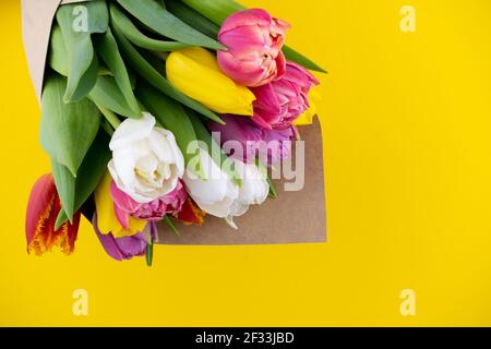 Ein Bouquet aus Tulpen, umhüllt von Bastelpapier. Frühlingsblumen. Geschenklieferung. Feiern. Speicherplatz kopieren. Gelbe, weiße, rosa, violette Knospen. Gelber Hintergrund. Postkarte. Tulpenblume Stockfoto