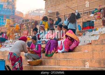17,12.2019, Varanasi, Indien. Einheimische verkaufen Schmuck, der in der Nähe der Treppe sitzt. Im Hintergrund Touristen mit Rucksäcken. Im Freien. Stockfoto