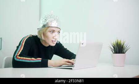 Ängstliche Frau mit Aluminium-Hut arbeiten auf Laptop. Schutz vor 5G und Gedankenlesen und Kontrolle. Hochwertige Fotos Stockfoto