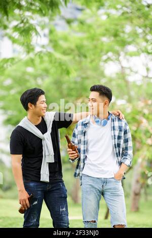 Zwei asiatische junge Männer trinken Bier und reden mit jedem Andere während ihres Spaziergangs im Park Stockfoto