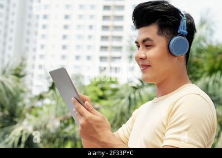 Asiatischer junger Mann mit Kopfhörern, der etwas online auf digital sieht Tablet in der Stadt Stockfoto
