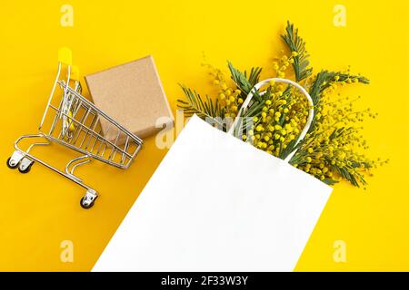 Ein Bouquet von Mimosen in einer weißen imitierten Papiertüte, Bastelschachtel und einem Lebensmittelkarren. Frühlingseinkäufe, Geschenke und Aktionen für International Women's D Stockfoto