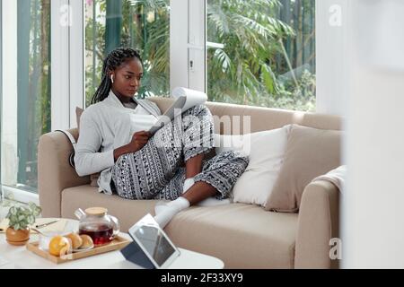 Konzentrierte schwarze junge Frau in Loungewear arbeitet von zu Hause aus, sitzt sie auf dem Sofa und liest Geschäftsdokument Stockfoto