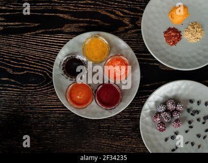 Auswahl an Saucen, Gewürzen, gefrorenen Beeren auf grauen Tellern auf einem dunklen Holztisch. Blick von oben. Stockfoto
