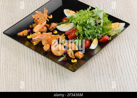 Salat mit Garnelen, Muscheln, Rucola, Tomaten, Eiern, Salat auf einem schwarzen quadratischen Teller auf einem weißen Holztisch. Stockfoto