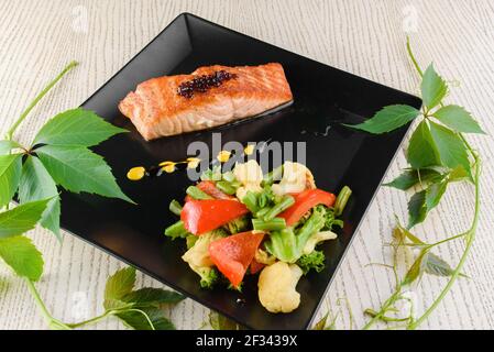 Lachsfilet mit gebratenem Gemüse auf quadratischen schwarzen Tellern Ein weißer Holztisch Stockfoto
