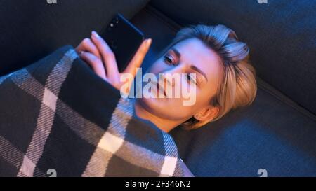 Portrait einer jungen Frau, die in den sozialen Medien unterwegs ist, während sie sich auf dem mit einer Decke bedeckten Sofa entspannt. Hochwertige Fotos Stockfoto