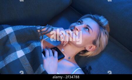 Junge blonde Frau SMS an ihren Freund, während sie sich auf der mit Decke bedeckten Couch entspannt. Hochwertige Fotos Stockfoto