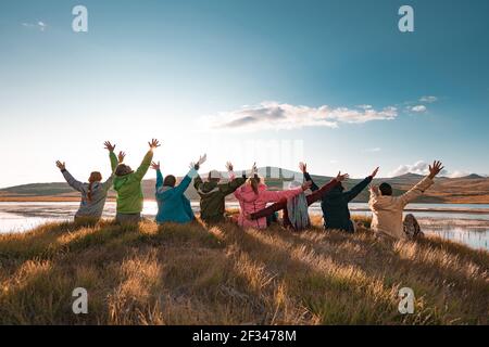 Große Gruppe von glücklichen Touristen sitzen mit erhobenen Armen Bei schönem Sonnenuntergang See und Berge Stockfoto