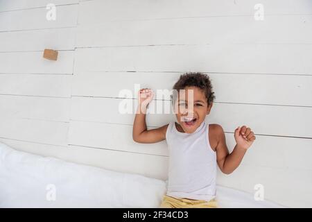 Kleiner afroamerikanischer Junge liegt auf dem Holzboden in weißem T-Shirt, gelbe Shorts. Kind spielt, hat Spaß, spielt im Zimmer am Bett. Co Stockfoto