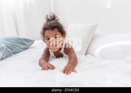 Kleine afroamerikanische Mädchen mit lockigen Haaren in Bun gebunden sitzt auf weißem Bett in Loft skandinavischen Interieur. Kleines Mädchen mit Spaß auf Sofa unter blau Stockfoto