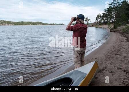 Der Mann steht an einem Sandstrand, am Ufer eines Sees und schaut mit Interesse durch Ferngläser, neben einem geparkten Kajak. Stockfoto