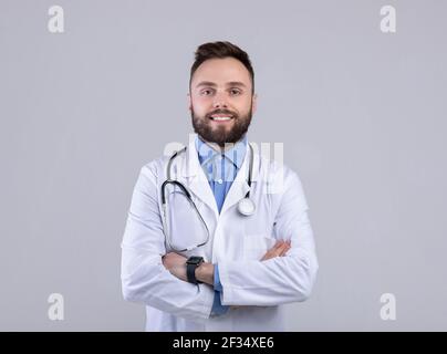 Porträt von positiven männlichen Arzt posiert mit gekreuzten Armen und Lächelnd auf Kamera über grauem Studiohintergrund Stockfoto