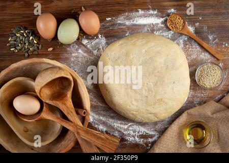 Teig zum Aufgehen übrig, Mehl; Holzgeräte, Eier und verschiedene Samen auf einem Walnussbrett Stockfoto