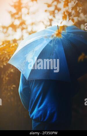 Ein Mann in einer blauen Jacke und mit einem blauen Regenschirm in der Hand geht allein im Park an einem sonnigen Herbsttag. Oktober. Stockfoto