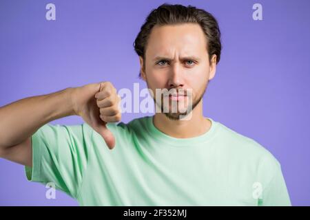 Junger gutaussehender Mann, der Unzufriedenheit ausdrückt und mit dem Daumen nach unten auf die Kamera zeigt. Kerl mit Bart stehen auf violettem Studio-Hintergrund. zeichen Stockfoto