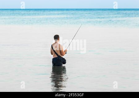 Ein junger Mann ohne Hemd steht am Abend knietief im Wasser des Meeres und fängt mit einer Leine Fische. Stockfoto