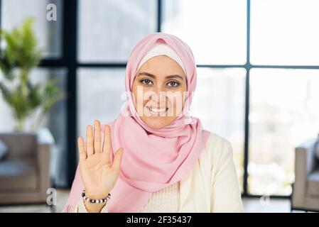Close shot Porträt von lächelnden muslimischen Frau tragen leichte Kleidung und rosa nationalen Hijab, posiert für Foto am Arbeitsplatz, glücklich aufgeregt, sitzen an der Kamera, winken die Hand, machen Videoanruf Stockfoto