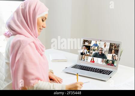 Arabische Frau in Hijab mit einem Video-Chat auf Laptop, im Gespräch mit der Web-Kamera, sitzen am Schreibtisch, schriftlich Arbeitsauftrag im Notebook, Online-Nachhilfe, Lehrer mit Videoanruf mit erwachsenen Schülern Stockfoto