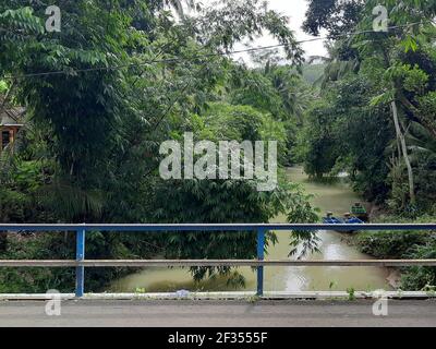 Ein grün brauner Fluss flankiert von tropischen Regenwald Bäumen in Gunung Kidul auf Java, Indonesien. Blick von einer Brücke mit Eisengeländer. Stockfoto