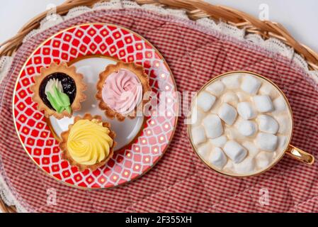 Leckere Cupcakes und Tasse Kakao mit Marshmallows auf Weidenschale, Vanillecupcakes mit rosa und gelber Sahne. Stockfoto