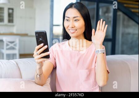 Lächelnde asiatische Frau mit weißen Zähnen und intelligente Uhr am Handgelenk, Blick auf das Telefon, zu Hause sitzen, im Gespräch mit Verwandten, winkende Hand Video Anruf Distanz Freund online in mobilen Chat-App Stockfoto
