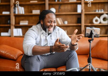 Blogging-Konzept. Junge afroamerikanische männliche Blogger in Freizeitkleidung Streaming-Video online oder Aufnahme Tutorial für Social Media-Netzwerk, Blick auf das Handy auf einem Stativ und lachen Stockfoto