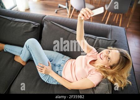 Fröhliche Frau liegt auf dem Sofa und macht Selfie zu Hause Stockfoto