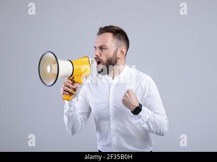 Aufgeregter junger Mann, der auf grauem Studiohintergrund in ein Megaphon schreit Stockfoto