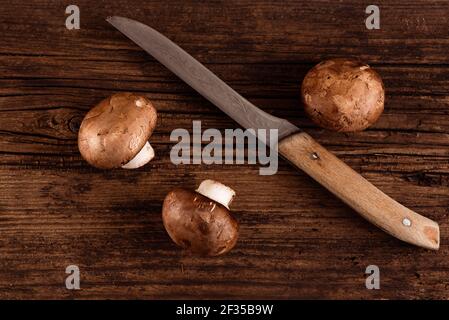 Cremini Pilze und altes Küchenmesser auf texturiertem Holzhintergrund. Drei Baby portobello. Draufsicht. Nahaufnahme Stockfoto