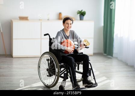 Paralympics-Champion. Fröhlicher Teenager Junge im Rollstuhl hält Basketball und Trophäe, glücklich über seinen Sieg zu Hause Stockfoto