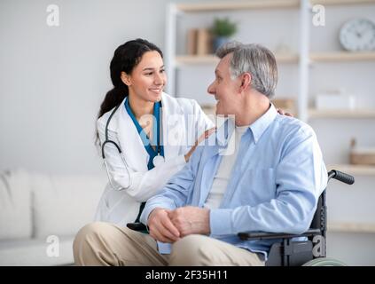 Behinderter älterer Mann im Rollstuhl und junge Krankenschwester, die sich um sie kümmert Von ihm zu Hause Stockfoto
