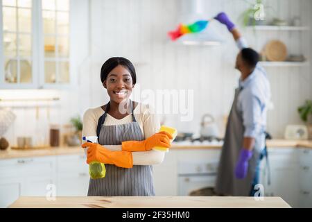 Porträt der schwarzen Dame in Schürze und Gummihandschuhe halten Waschmittel, lächelnd an der Kamera, Freund Staub auf dem Hintergrund Stockfoto