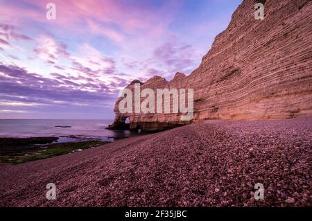 Hohe und steile erodierte Kreidefelsen an der Küste in der Dämmerung. Rosafarbener Himmel über dem Felsbogen Porte d'Amont, Etretat, Frankreich. Stockfoto