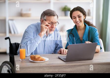 Junge Krankenschwester zeigt älteren behinderten Mann, wie man Laptop zu verwenden Computer im Altersheim Stockfoto