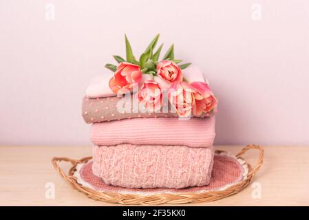 Stapel von gefalteten Wollgestrickpulvern oder Pullovern in rosa Pastellfarben auf Tisch mit Blumenstrauß Tulpen. Nahaufnahme von warmen gemütlich bequem monochon Stockfoto
