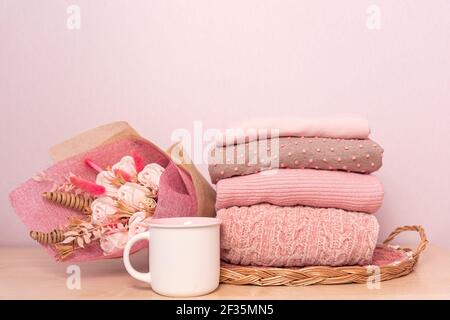 Stapel von gefalteten Wollgestrickpulvern oder Pullovern in rosa Pastellfarben und mag auf Tisch mit Blumenstrauß. Nahaufnahme von warmen gemütlichen bequemen Mono Stockfoto