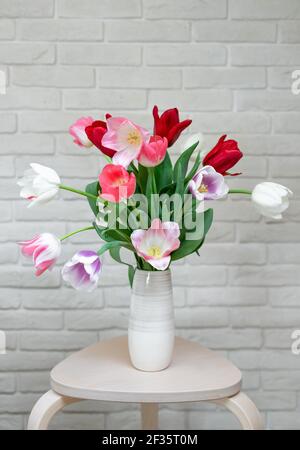 Bunte bunte bunte Tulpen in einer Vase in einem modernen Interieur gegen eine weiße Ziegelwand. Frühlingskarte. Vertikales Bild. Stockfoto