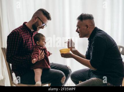 Männlich Gay paar mit angenommen Baby Mädchen zu Hause - Zwei schöne Väter füttern das Baby Mädchen in der Küche - Männliche Babysitter - LGBT Familie zu Hause - Vielfalt Stockfoto