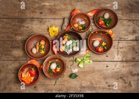 Osterschalen aus Keramik mit Eiern, Hasen und frischen Rosen auf einem Vintage-Tisch. Auswahl an traditionellen Gerichten mit frischen Blumen und festlicher Einrichtung. W Stockfoto