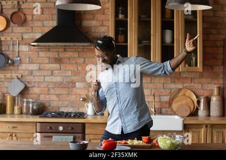 Fröhlicher afro amerikanischer Mann, der in der Küche mit improvisiertem Mikrofon singt Stockfoto
