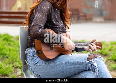 Kaukasische Mädchen spielen Gitarre im Freien sitzen auf einem Stuhl. In Jeans und einem legeren Hemd und mit ihrem Handy in der Gesäßtasche ihres tr Stockfoto