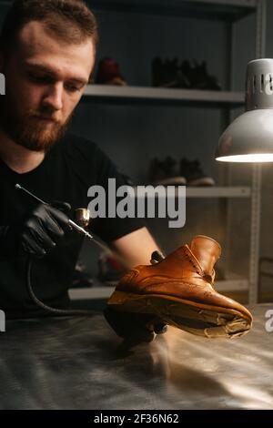 Nahaufnahme Vertikale Aufnahme eines bärtigen Schusters mit schwarzen Handschuhen, der Farbe von hellbraunen Lederschuhen sprüht, Nahaufnahme. Stockfoto