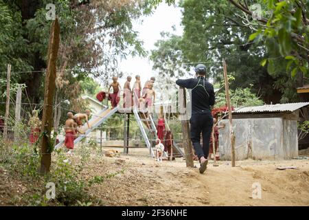 BAGAN, NYAUNG-U, MYANMAR - 2. JANUAR 2020: Ein Tourist steht und beobachtet mehrere junge Mönche spielen und Spaß haben auf einem lokalen Spielplatz mit einer Rutsche Stockfoto