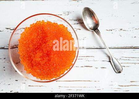 Eine transparente Schüssel mit rotem Lachskaviar und einem Silber Löffel auf einem weißen Holztisch Stockfoto