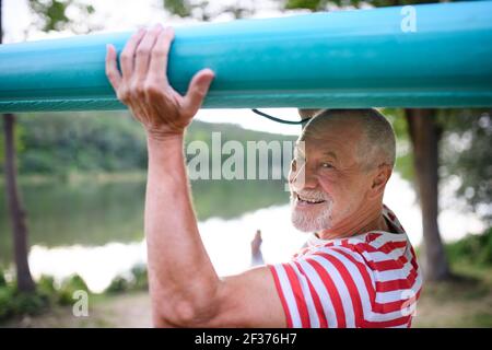 Älterer Mann, der im Sommer das Paddleboard am See trägt und die Kamera anschaut. Stockfoto