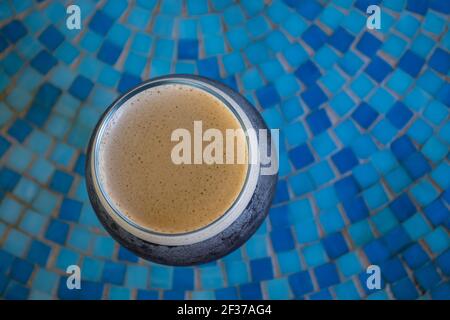 Rundes Glas kaltgebrühten Kaffees mit Schaumstoffplatte sitzend Auf einem blauen Mosaik gefliesten Tisch in einem Café im Freien Stockfoto